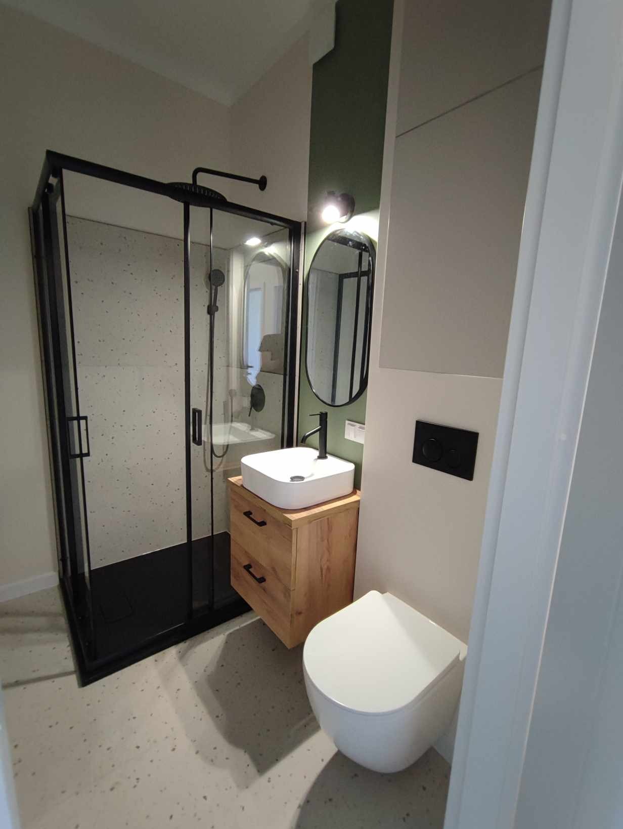 Nowoczesna łazienka z czarną kabiną prysznicową, umywalką na drewnianej szafce z szufladami i owalnym lustrem z czarną ramą, na tle zielonej ściany.