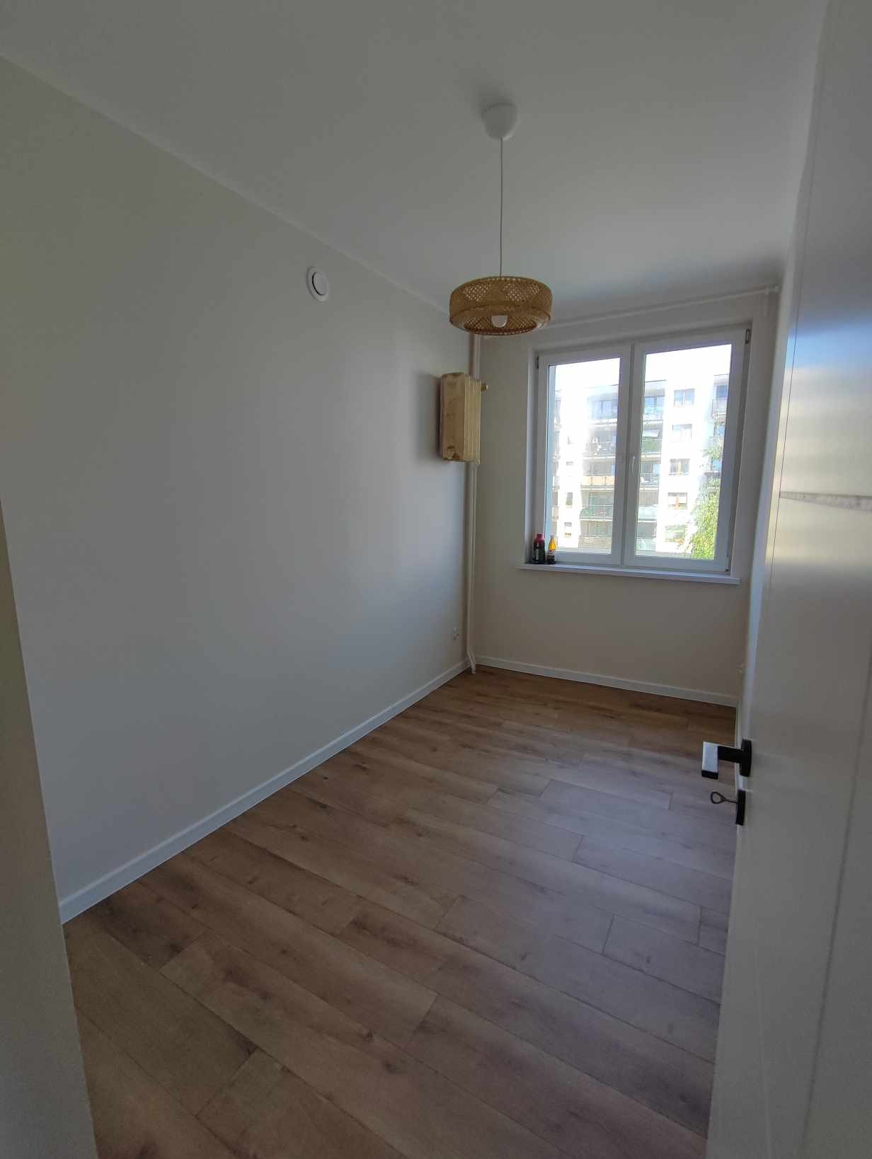 Pusty pokój po remoncie z drewnopodobnymi panelami podłogowymi, pomalowanymi na biało ścianami, okrągłą lampą z wikliny i grzejnikiem na ścianie.