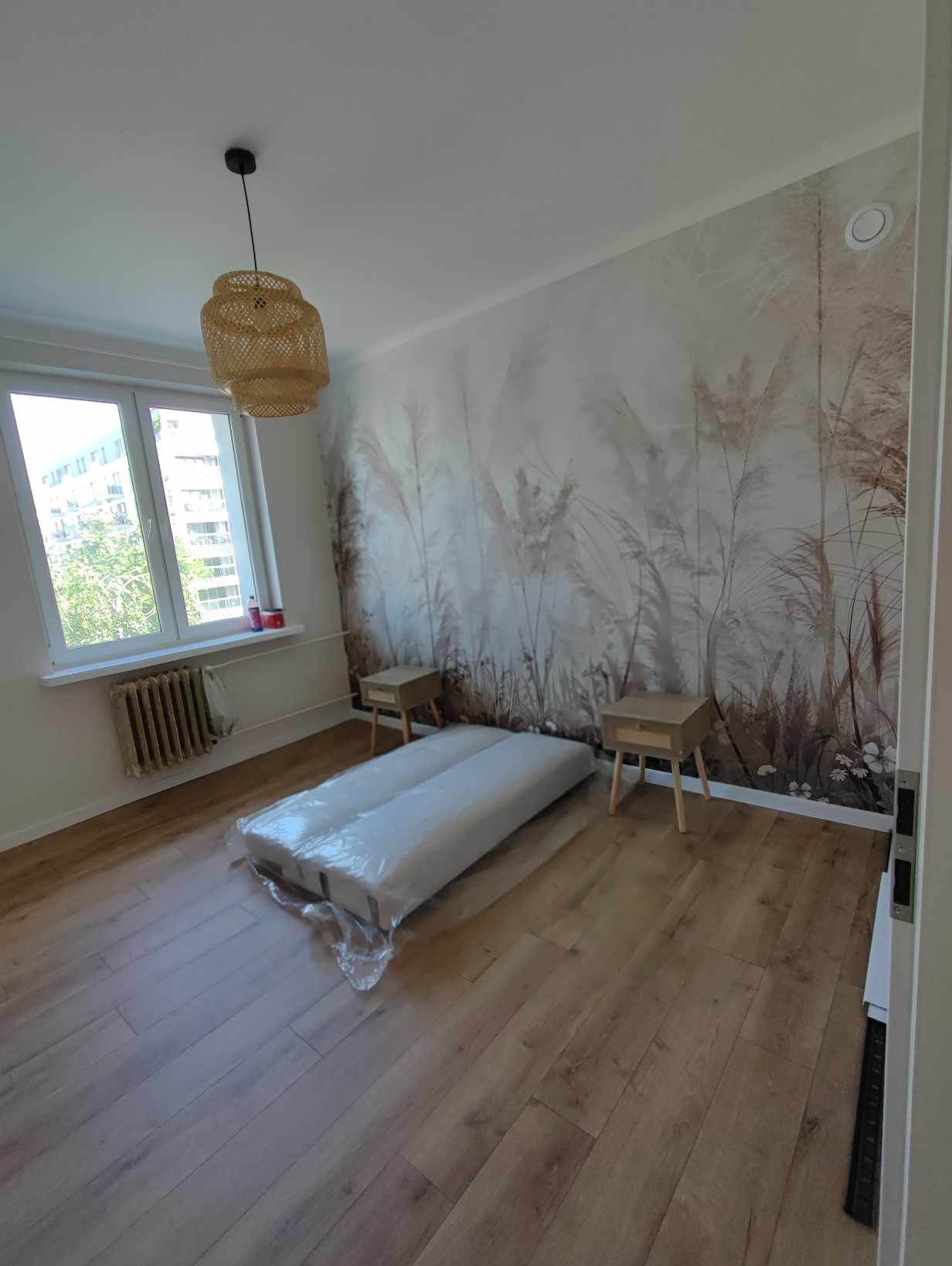Pokój po remoncie z nową podłogą z paneli, widoczny materac w folii, wzorzysta tapeta na ścianie i lampa z wikliny.