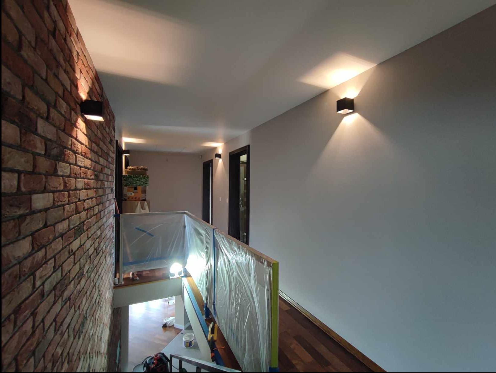 Korytarz w trakcie prac wykończeniowych: ściana z cegły, zabezpieczone folią schody, pomalowana szara ściana z nowoczesnymi kinkietami.