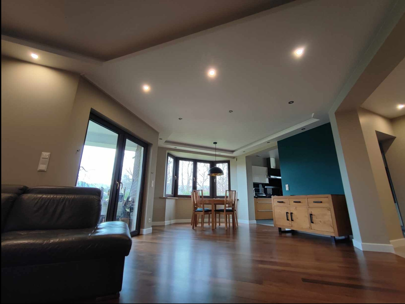 Przestronne wnętrze domu z drewnianą podłogą, nowoczesnym oświetleniem sufitowym, częściowo widoczną kuchnią i ciemnozieloną ścianą akcentującą, sfotografowane z niskiego kąta.