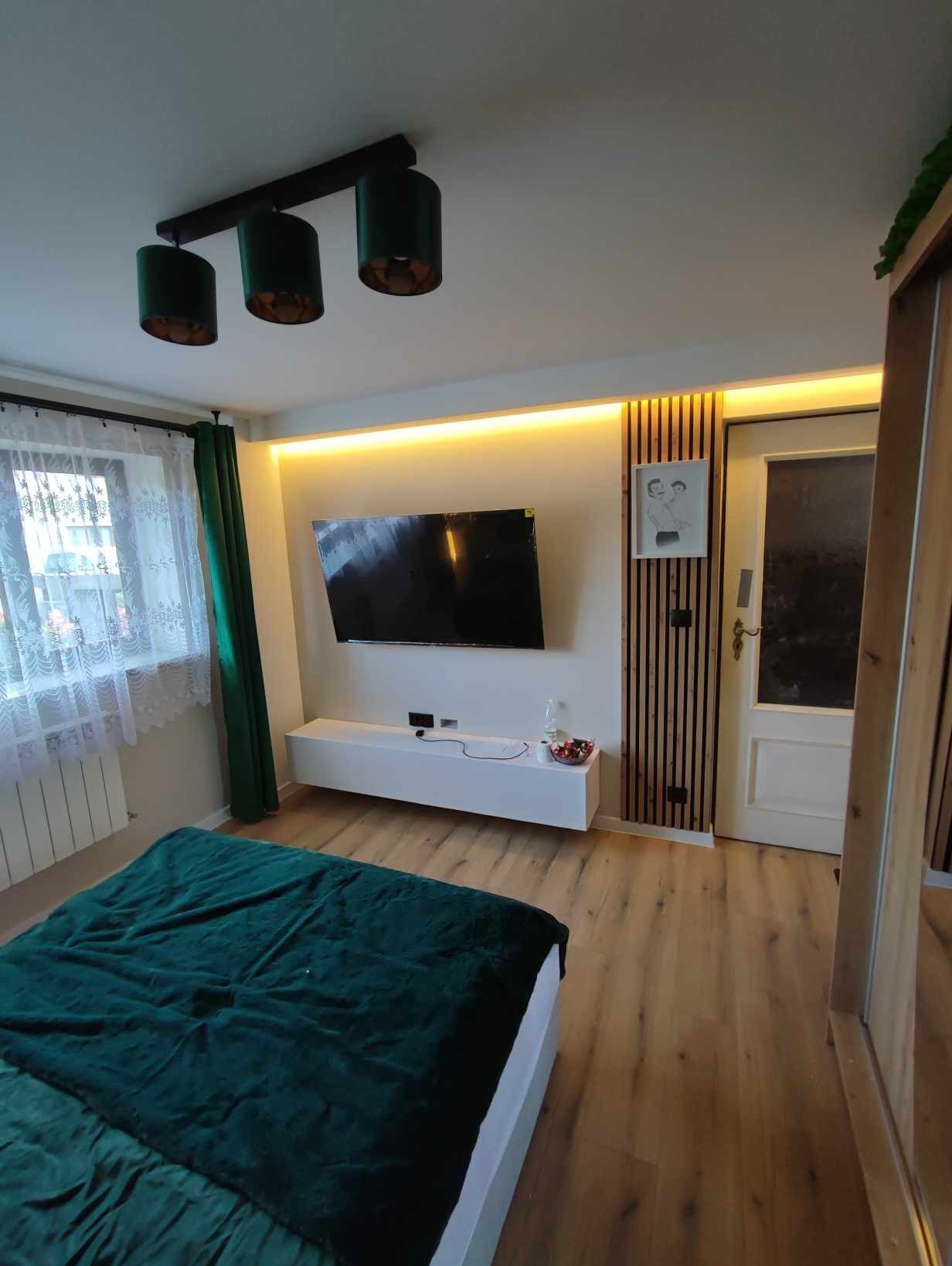 Nowoczesna sypialnia z podświetleniem LED za telewizorem, drewnianymi lamelami ściennymi i zielonymi akcentami w postaci zasłon i narzuty. Na ścianie wisi czarny telewizor, a pod nim biała szafka RTV.