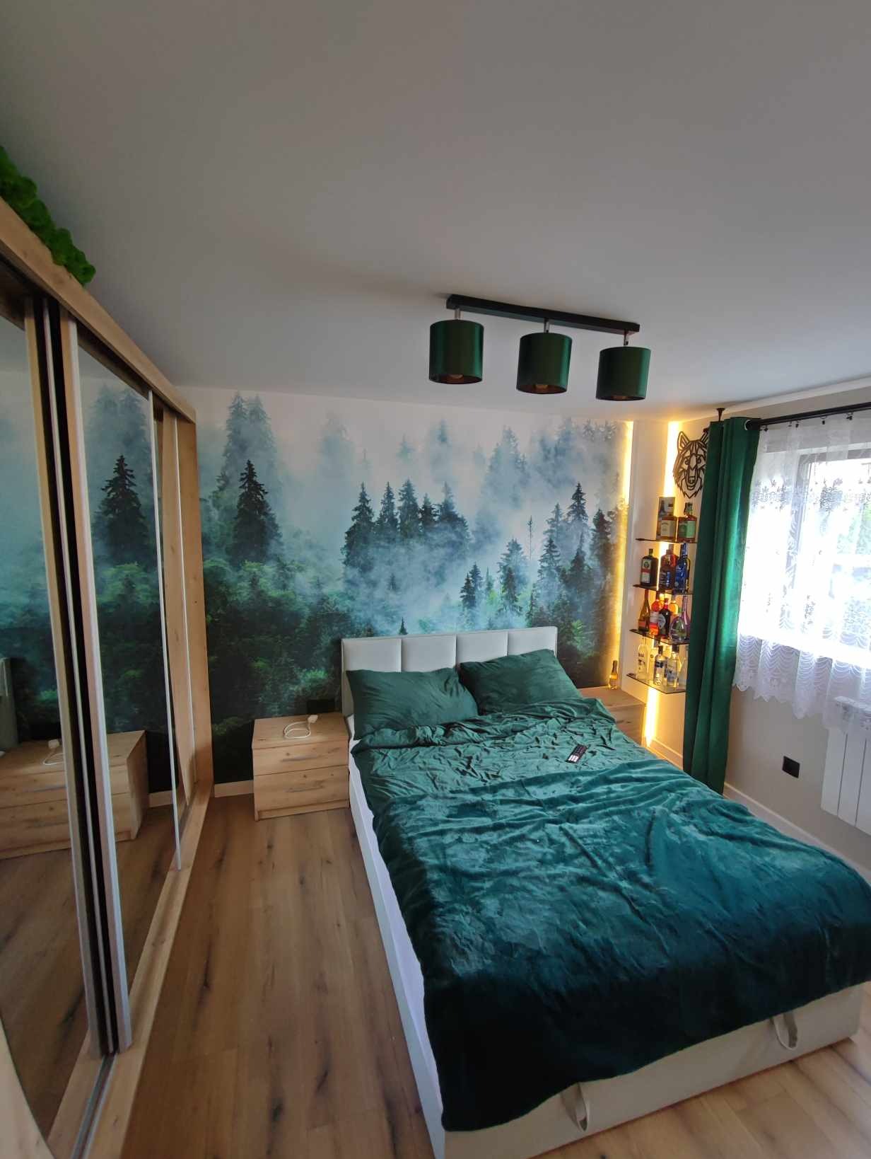 Sypialnia z fototapetą leśną, drewnianą podłogą i szafą z lustrami, oświetlona trzema zielonymi lampami i ledowym paskiem za półką z alkoholem.