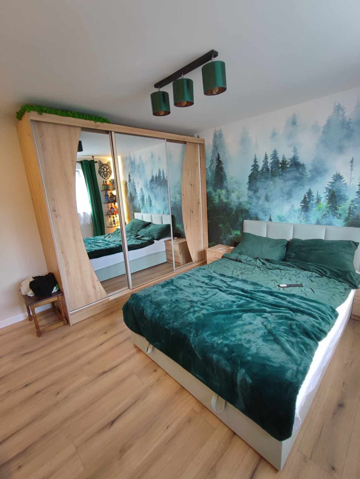 Sypialnia z łóżkiem w zielonej tonacji, tapetą z motywem lasu i dużą szafą z lustrami, na której umieszczono dekoracyjną zieleń.