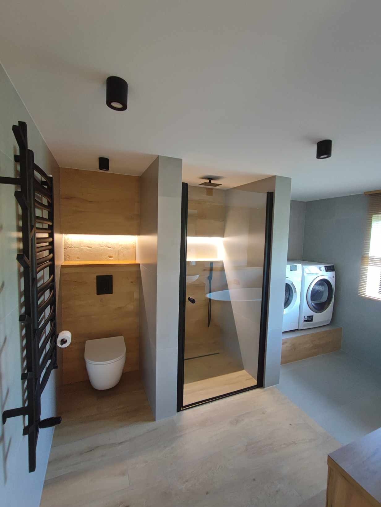 Nowoczesna łazienka z kabiną prysznicową typu walk-in, podświetlaną niszą z toaletą, grzejnikiem drabinkowym i pralką na podwyższeniu, wykończona w odcieniach szarości i drewna.