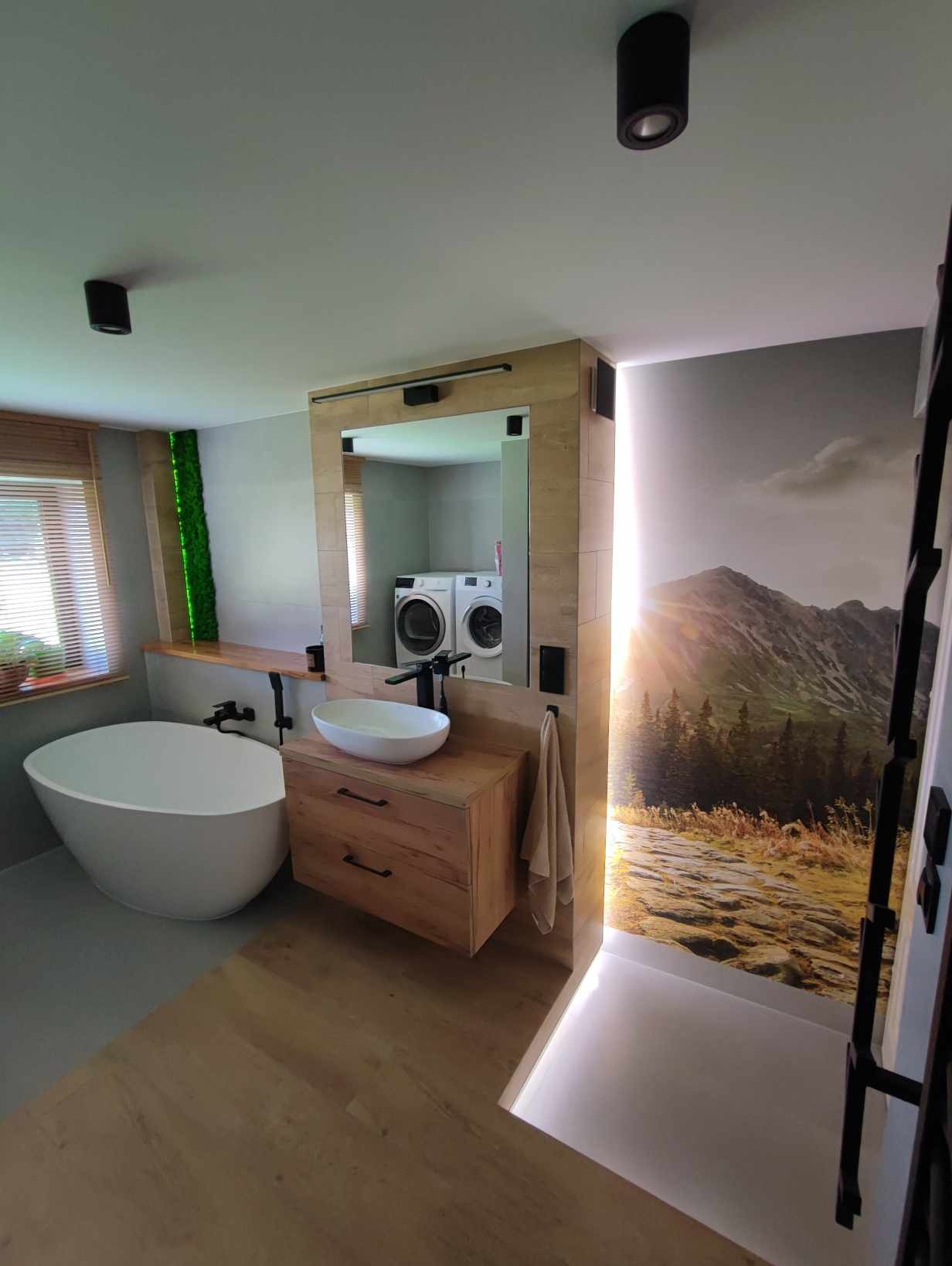 Nowoczesna łazienka z wolnostojącą wanną, umywalką nablatową na drewnianej szafce, lustrem z podświetleniem i fototapetą górską, w odbiciu lustra widoczne pralka i suszarka.