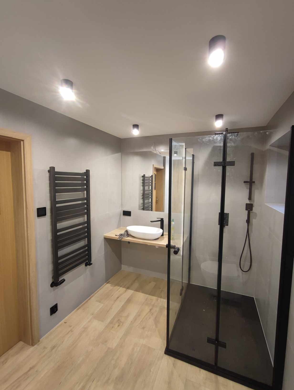 Nowoczesna łazienka z kabiną prysznicową typu walk-in z czarnymi profilami, umywalką nablatową na drewnianym blacie, grzejnikiem drabinkowym i panelami podłogowymi imitującymi drewno.