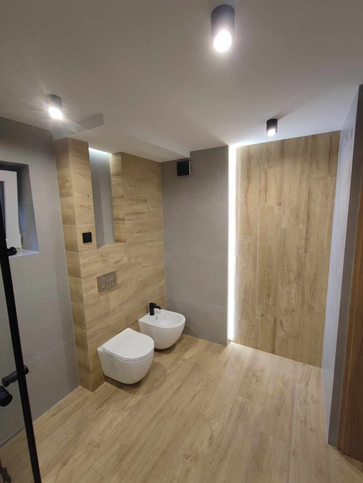 Nowoczesna łazienka z podwieszaną toaletą i bidetem, wykończona płytkami imitującymi drewno i szarymi panelami, oświetlona punktowymi lampami sufitowymi i pionową listwą LED.