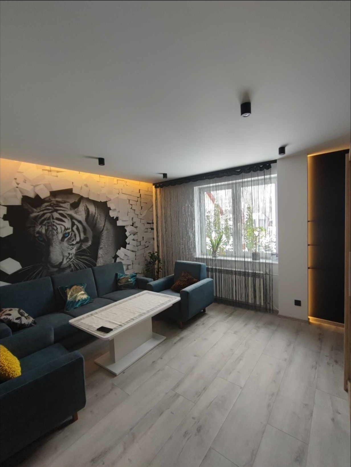 Salon z szarą podłogą z paneli, narożnikiem w odcieniach niebieskiego, fototapetą z motywem tygrysa i oświetleniem LED nad nią.