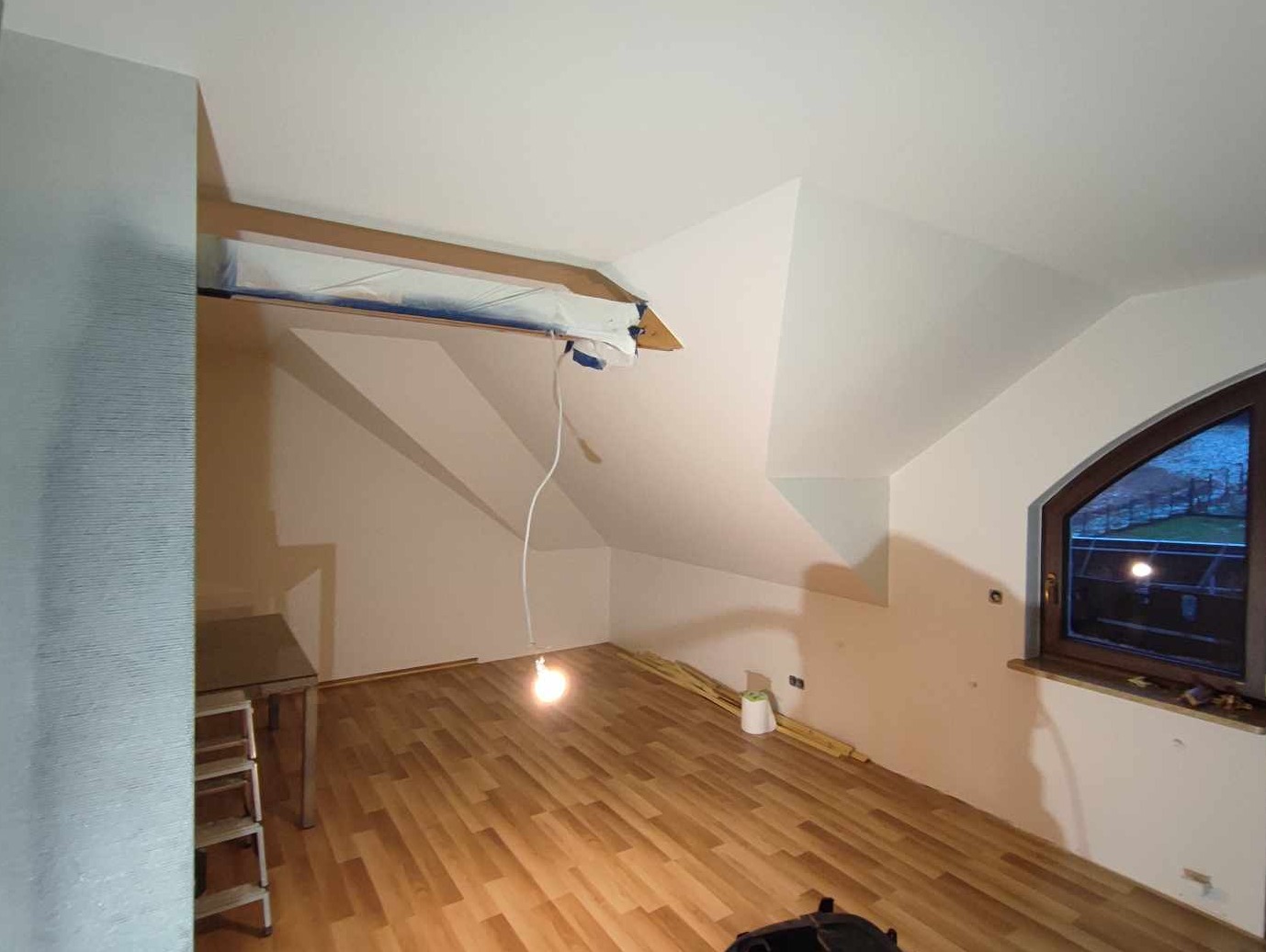 Pokój na poddaszu w trakcie przygotowań do malowania: zabezpieczona taśmą krawędź sufitu, widoczne fragmenty listew przypodłogowych, drewniana podłoga panelowa i wisząca żarówka.