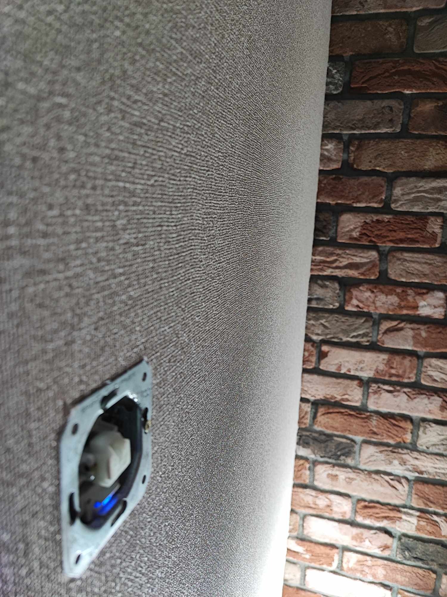 Szara, teksturowana ściana z zamontowanym gniazdkiem elektrycznym, sąsiadująca z fragmentem dekoracyjnej ściany z cegły.