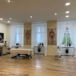 Jasne wnętrze gabinetu z łóżkiem do masażu przykrytym beżowym pledem, anatomiczne plansze wiszące na ścianach, drewniana podłoga i liczne rośliny doniczkowe.