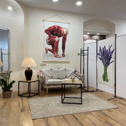 J&M Gabinet Masażu - Jasne wnętrze gabinetu z plakatem przedstawiającym anatomię mięśni, kanapą przykrytą wzorzystą narzutą, stolikami z lampkami, lustrem w ozdobnej ramie i parawanem z motywem lawendy.