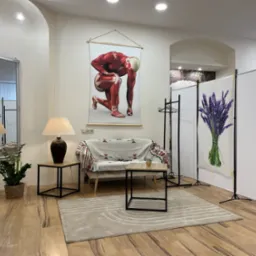 Jasne wnętrze gabinetu z plakatem przedstawiającym anatomię mięśni, kanapą przykrytą wzorzystą narzutą, stolikami z lampkami, lustrem w ozdobnej ramie i parawanem z motywem lawendy.