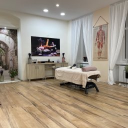 J&M Gabinet Masażu - Jasne wnętrze gabinetu z łóżkiem do masażu, dekoracjami z lawendy, plakatem anatomicznym i fototapetą z motywem toskańskiej uliczki.