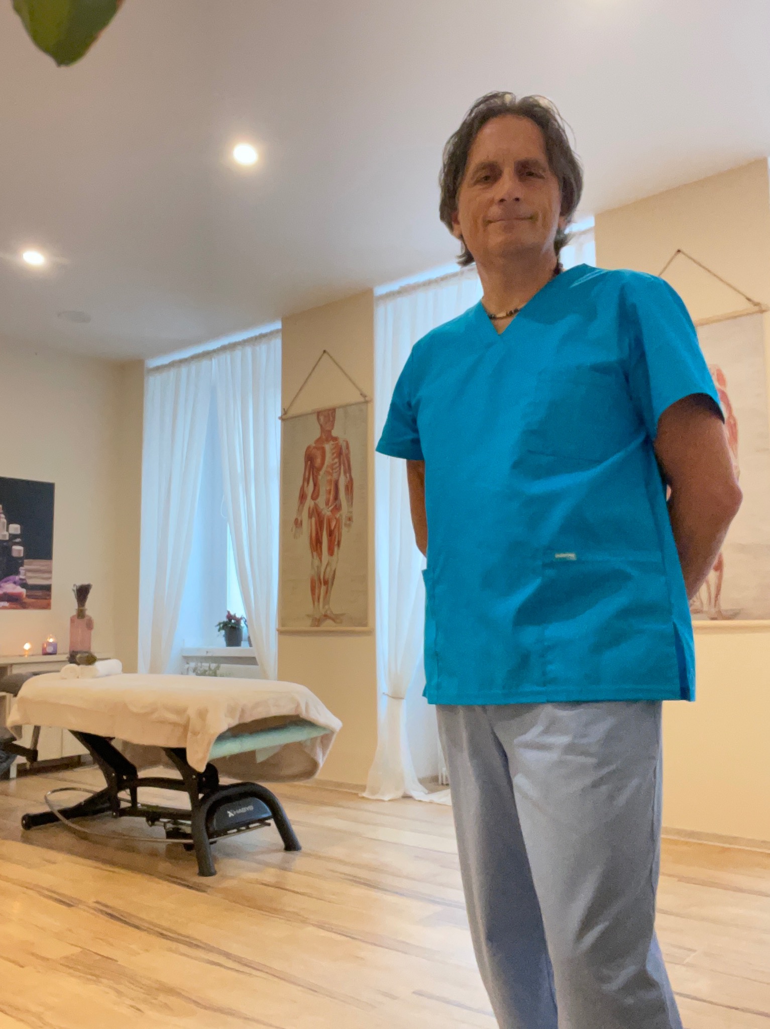 Uśmiechnięty mężczyzna w turkusowym uniformie stoi w gabinecie z kozetką do zabiegów i plakatem anatomicznym na ścianie.