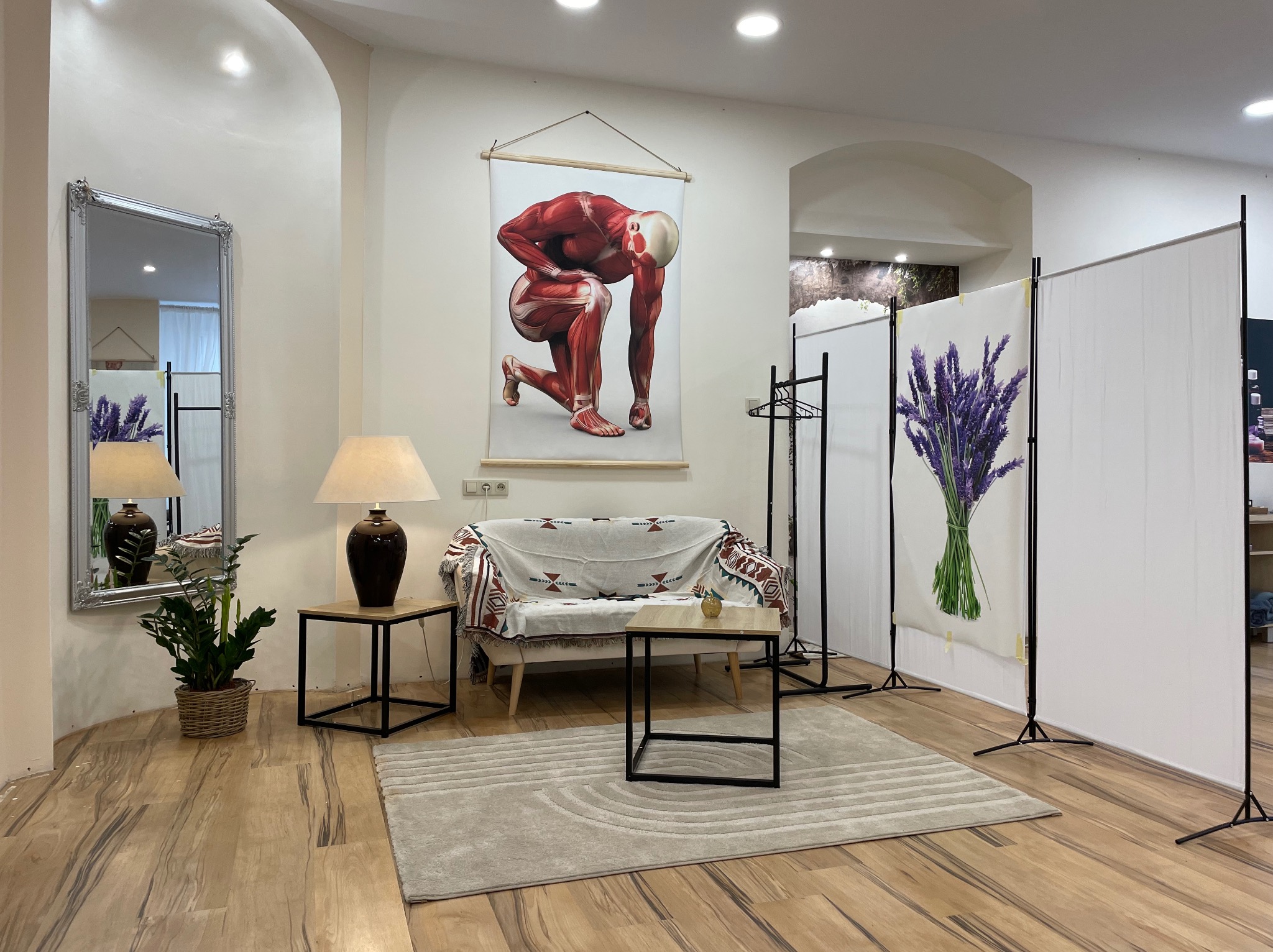 Jasne wnętrze gabinetu z plakatem przedstawiającym anatomię mięśni, kanapą przykrytą wzorzystą narzutą, stolikami z lampkami, lustrem w ozdobnej ramie i parawanem z motywem lawendy.