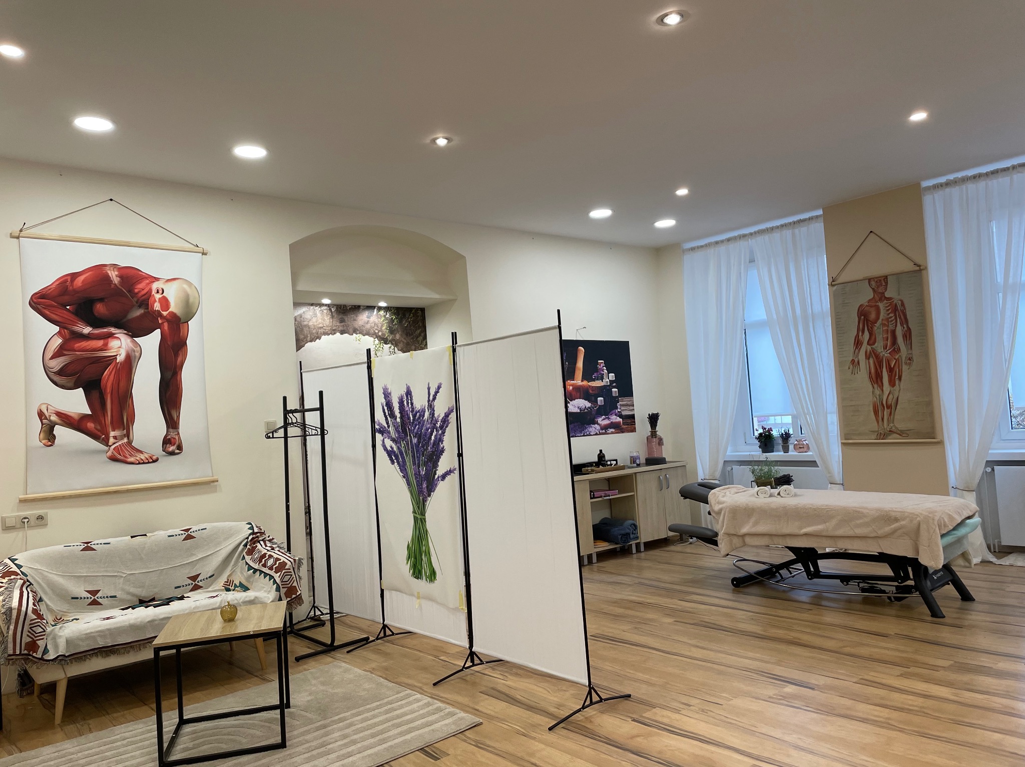 Jasne wnętrze gabinetu z kozetką do masażu przykrytą beżowym ręcznikiem, parawanem z motywem lawendy, grafikami anatomicznymi na ścianach i sofą z wzorzystą narzutą.