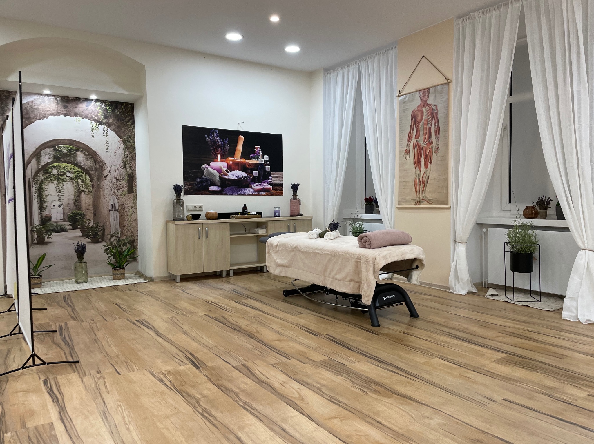 Jasne wnętrze gabinetu z łóżkiem do masażu, dekoracjami z lawendy, plakatem anatomicznym i fototapetą z motywem toskańskiej uliczki.