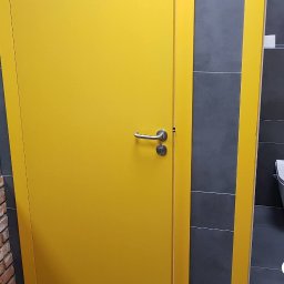 Ewa Musialik-Sojka Out-In - Wnętrze toalety publicznej z żółtymi drzwiami kabiny i szarą glazurą, widoczna muszla klozetowa i fragment ściany z cegły.