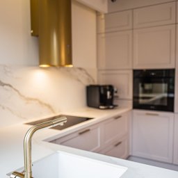 Ewa Musialik-Sojka Out-In - Szczegółowe ujęcie nowoczesnej kuchni z białym marmurowym blatem, złotą baterią i cylindrycznym okapem w kolorze złota, zintegrowaną płytą grzewczą oraz zabudowaną szafką z piekarnikiem i ekspresem...