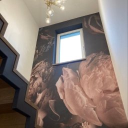 Ewa Musialik-Sojka Out-In - Klatka schodowa z ciemną tapetą w kwiaty, białe okno na ścianie, złoty żyrandol z kulistymi kloszami i drewniane schody z geometryczną balustradą.