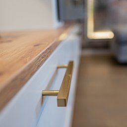 Ewa Musialik-Sojka Out-In - Biała szafka z drewnianym blatem i złotym, kwadratowym uchwytem, widoczna z bliska pod kątem.
