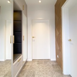 Ewa Musialik-Sojka Out-In - Nowoczesny przedpokój z szafą wnękową z lustrami, siedziskiem z czarną tapicerką i złotymi uchwytami, wzorzystą podłogą oraz drewnianą okładziną ściany w jodełkę.