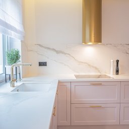 Ewa Musialik-Sojka Out-In - Nowoczesna kuchnia z pudrowo-różowymi szafkami, białym blatem imitującym marmur i złotym okapem cylindrycznym, uzupełniona o minimalistyczną armaturę i zioła w doniczce przy oknie.