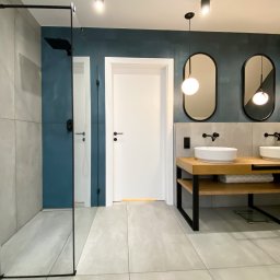 Ewa Musialik-Sojka Out-In - Nowoczesna łazienka z kabiną prysznicową typu walk-in, podwójną umywalką na drewnianym blacie z czarnymi nogami, owalnymi lustrami podwieszonymi na czarnych przewodach oraz niebiesko-szarą ścianą.