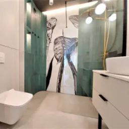 Nowoczesna łazienka z podwieszaną toaletą, białą szafką z umywalką nablatową i szklaną kabiną prysznicową z motywem liści na ścianie.