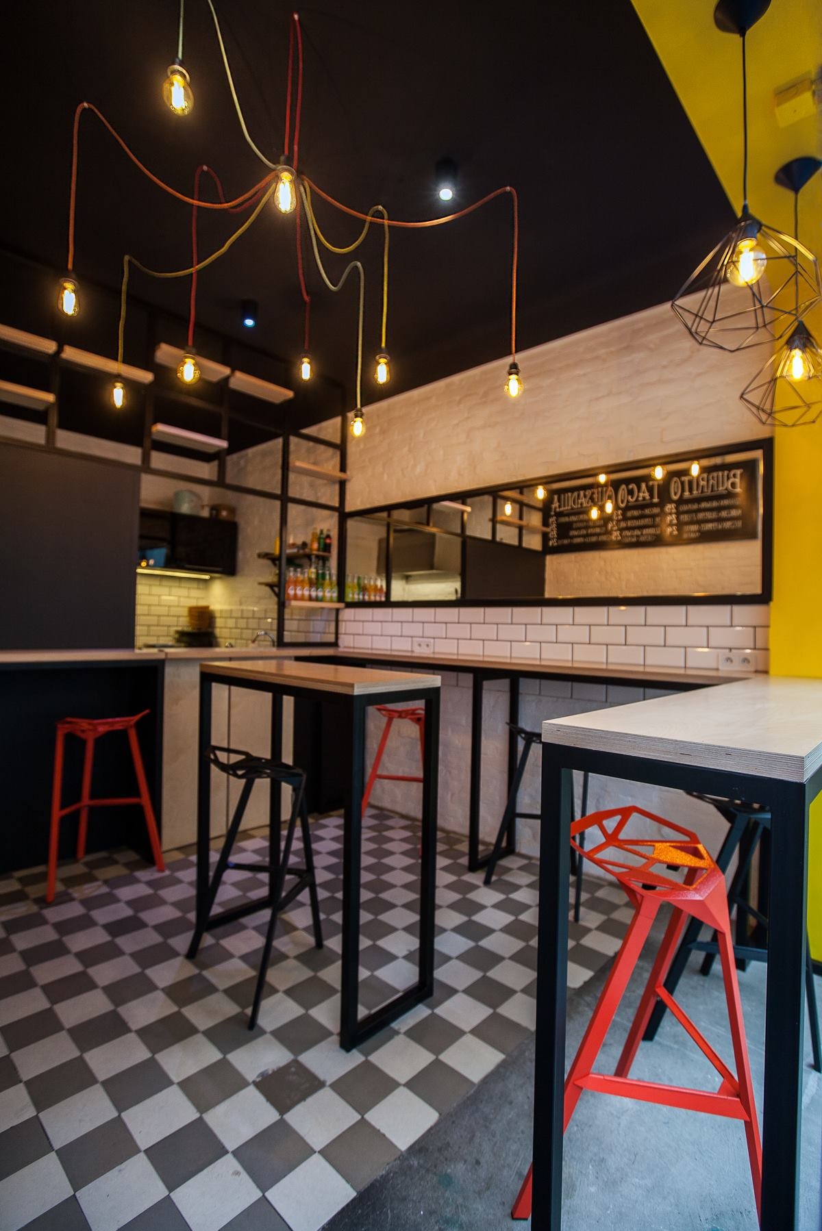 Wnętrze lokalu gastronomicznego z blatem barowym, krzesłami o geometrycznej konstrukcji w kolorze czerwonym i czarnym, oraz oświetleniem w stylu industrialnym z żarówkami Edisona i wzorzystą...