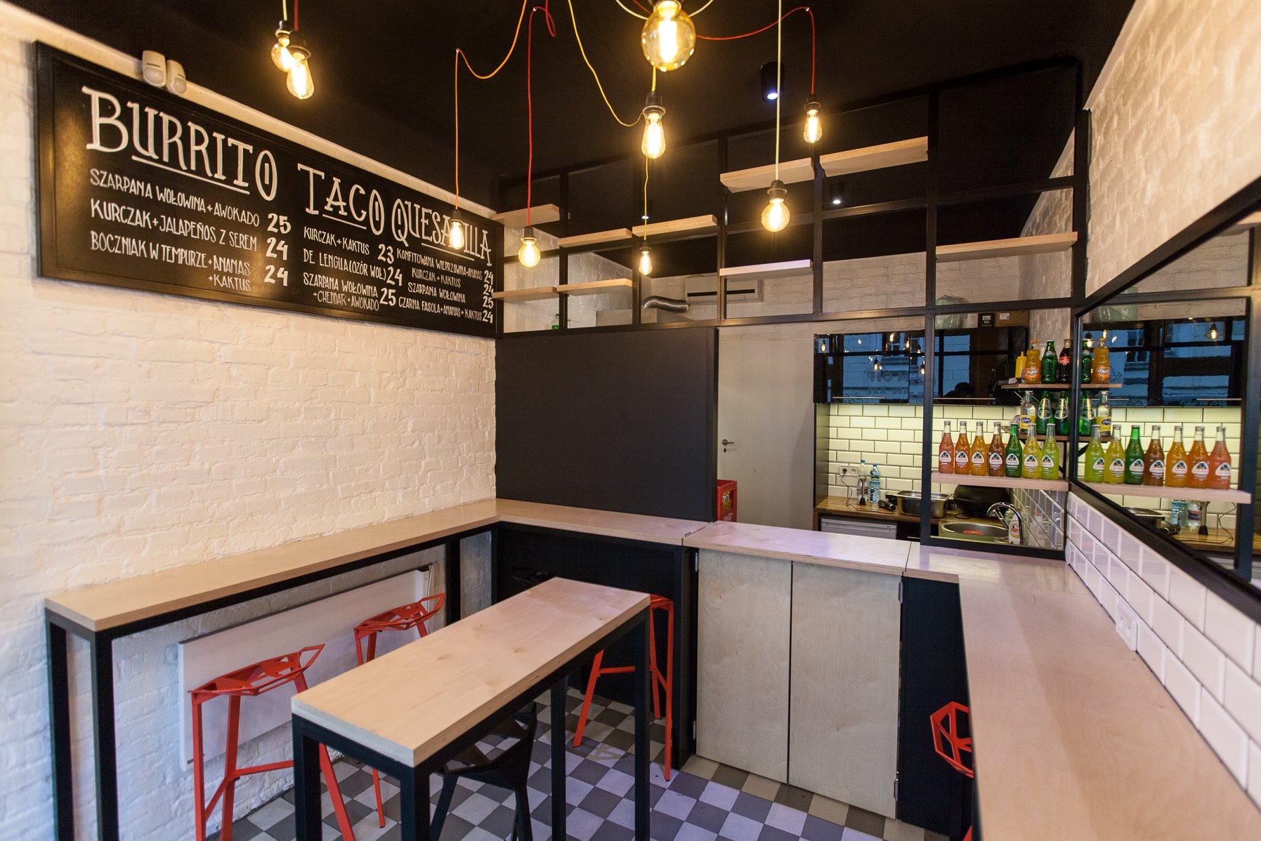 Wnętrze lokalu gastronomicznego w stylu loftowym z menu wypisanym kredą na czarnej tablicy, wiszące żarówki edisona, białe cegły na ścianach i szaro-biała szachownica na podłodze.