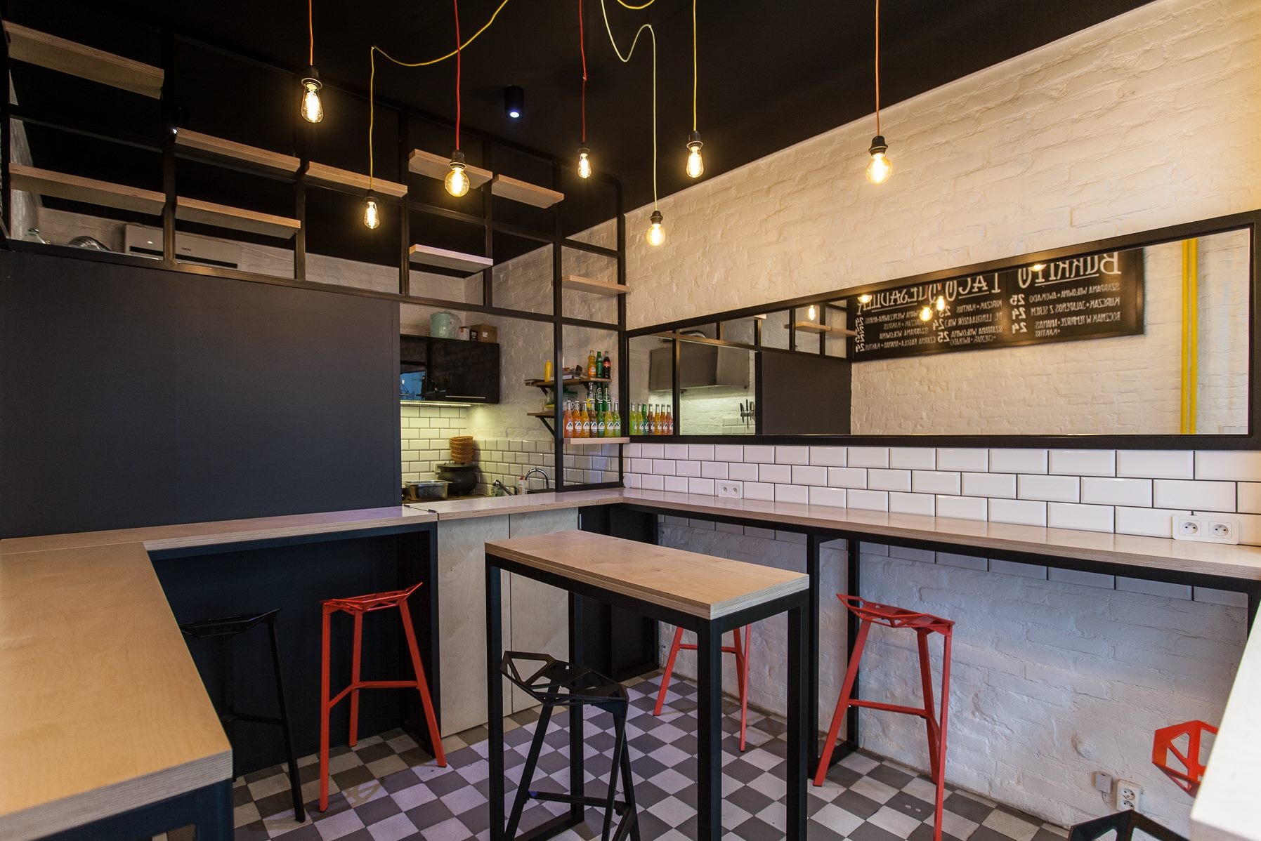 Nowoczesne wnętrze lokalu gastronomicznego z ceglanymi ścianami, czarnym sufitem z wiszącymi żarówkami, wysokimi stolikami barowymi i kolorowymi stołkami, szachownicą na podłodze oraz lustrem z menu.