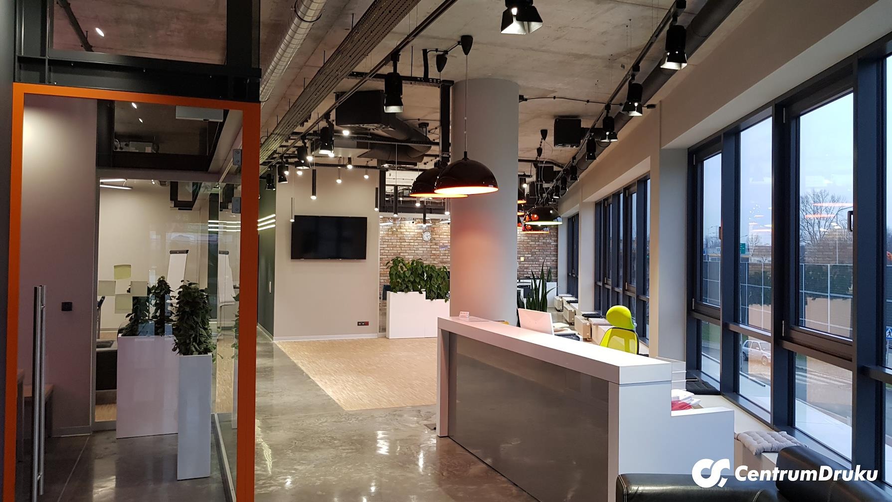 Nowoczesne biuro z recepcją, widok na przeszklone pomieszczenie z roślinami, surowy betonowy sufit z widocznymi instalacjami, lampy wiszące nad recepcją, ściana z cegły w tle.