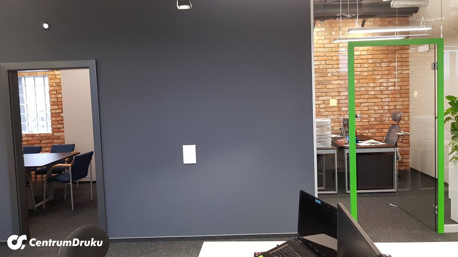 Nowoczesne biuro z szarą ścianą, widok na salę konferencyjną z ceglaną ścianą i przeszklone biuro z zielonymi ramami, widoczne biurko i roślina.