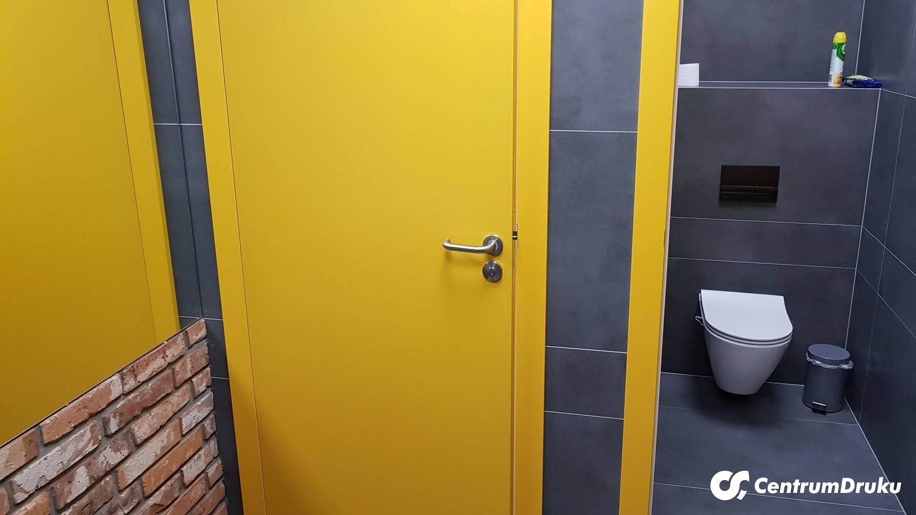Wnętrze toalety publicznej z żółtymi drzwiami kabiny i szarą glazurą, widoczna muszla klozetowa i fragment ściany z cegły.