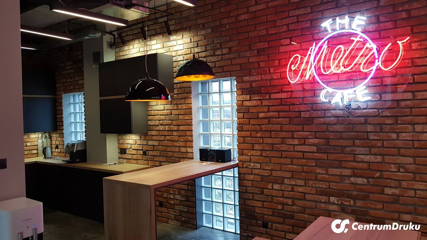 Nowoczesna aranżacja wnętrza biura z ceglaną ścianą, neonem 'The Metro Cafe', czarnymi lampami i aneksem kuchennym z drewnianym blatem.