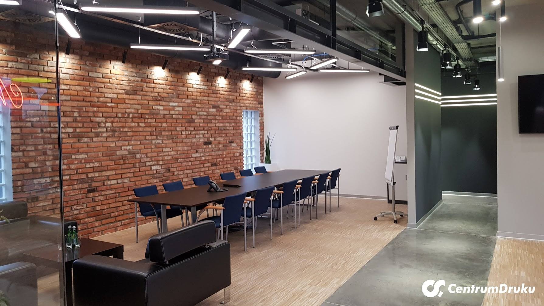 Sala konferencyjna w biurze w Warszawie z długim stołem, krzesłami w kolorze granatowym i ścianą z czerwonej cegły, oświetlona nowoczesnymi lampami sufitowymi.