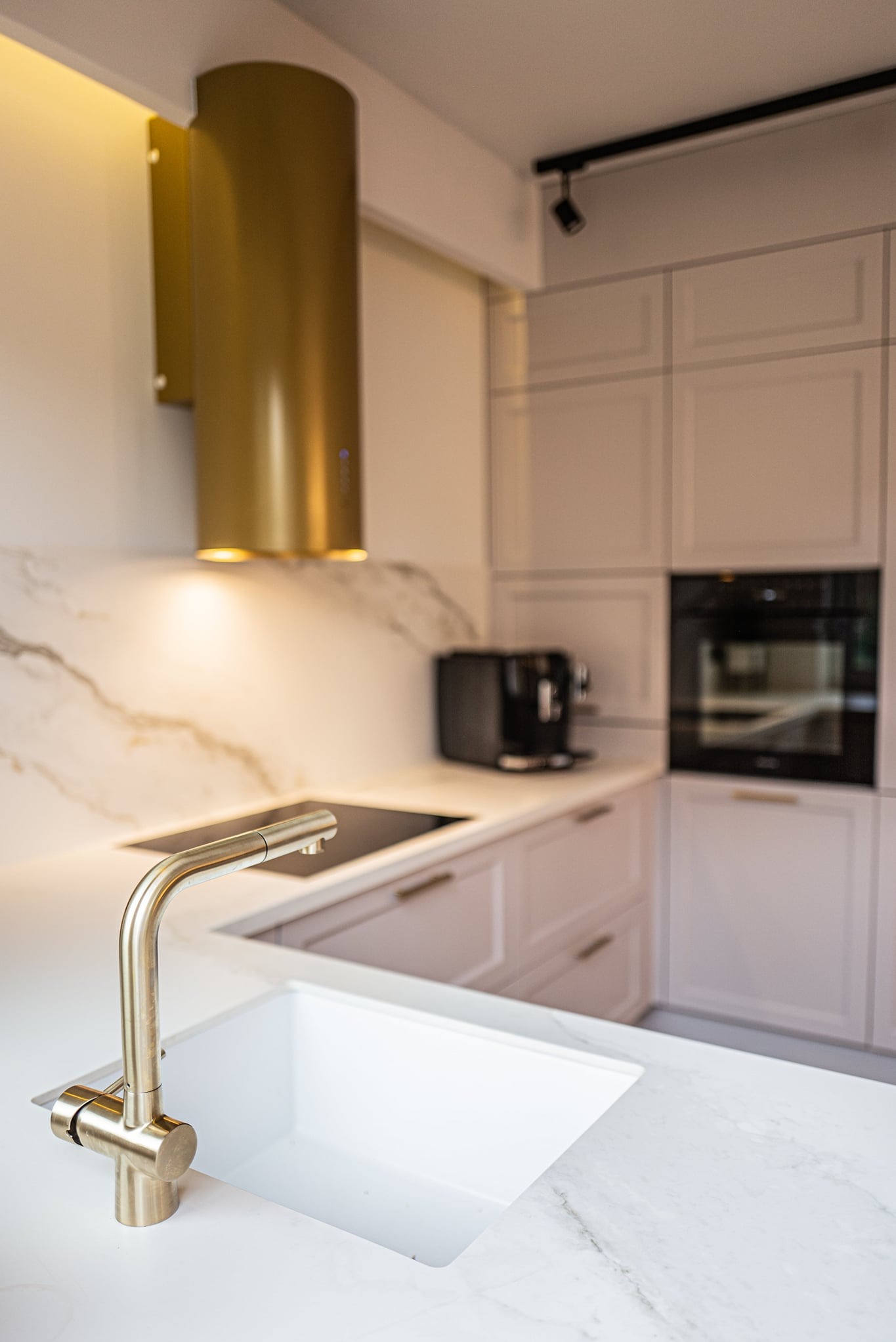 Szczegółowe ujęcie nowoczesnej kuchni z białym marmurowym blatem, złotą baterią i cylindrycznym okapem w kolorze złota, zintegrowaną płytą grzewczą oraz zabudowaną szafką z piekarnikiem i ekspresem...