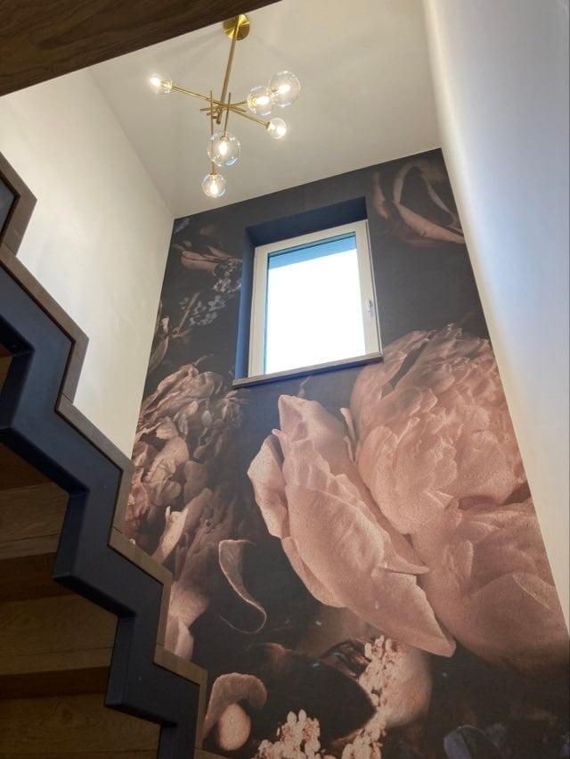 Klatka schodowa z ciemną tapetą w kwiaty, białe okno na ścianie, złoty żyrandol z kulistymi kloszami i drewniane schody z geometryczną balustradą.