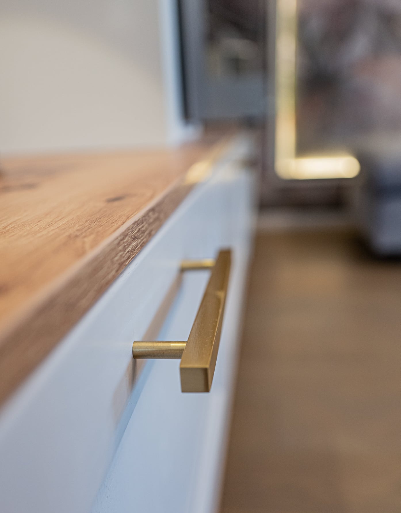 Biała szafka z drewnianym blatem i złotym, kwadratowym uchwytem, widoczna z bliska pod kątem.
