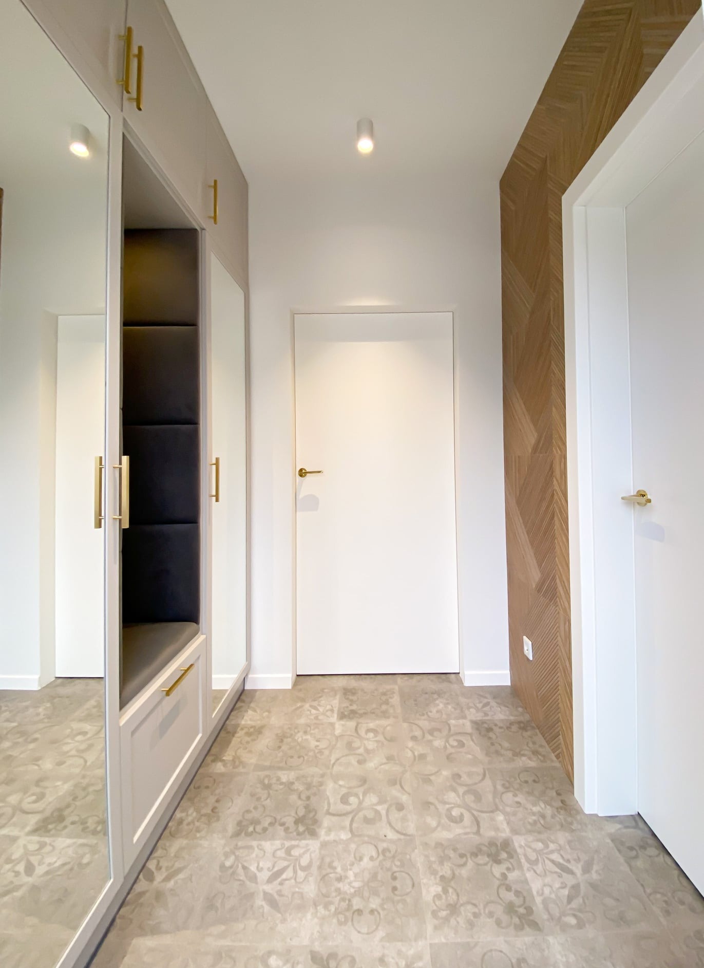 Nowoczesny przedpokój z szafą wnękową z lustrami, siedziskiem z czarną tapicerką i złotymi uchwytami, wzorzystą podłogą oraz drewnianą okładziną ściany w jodełkę.