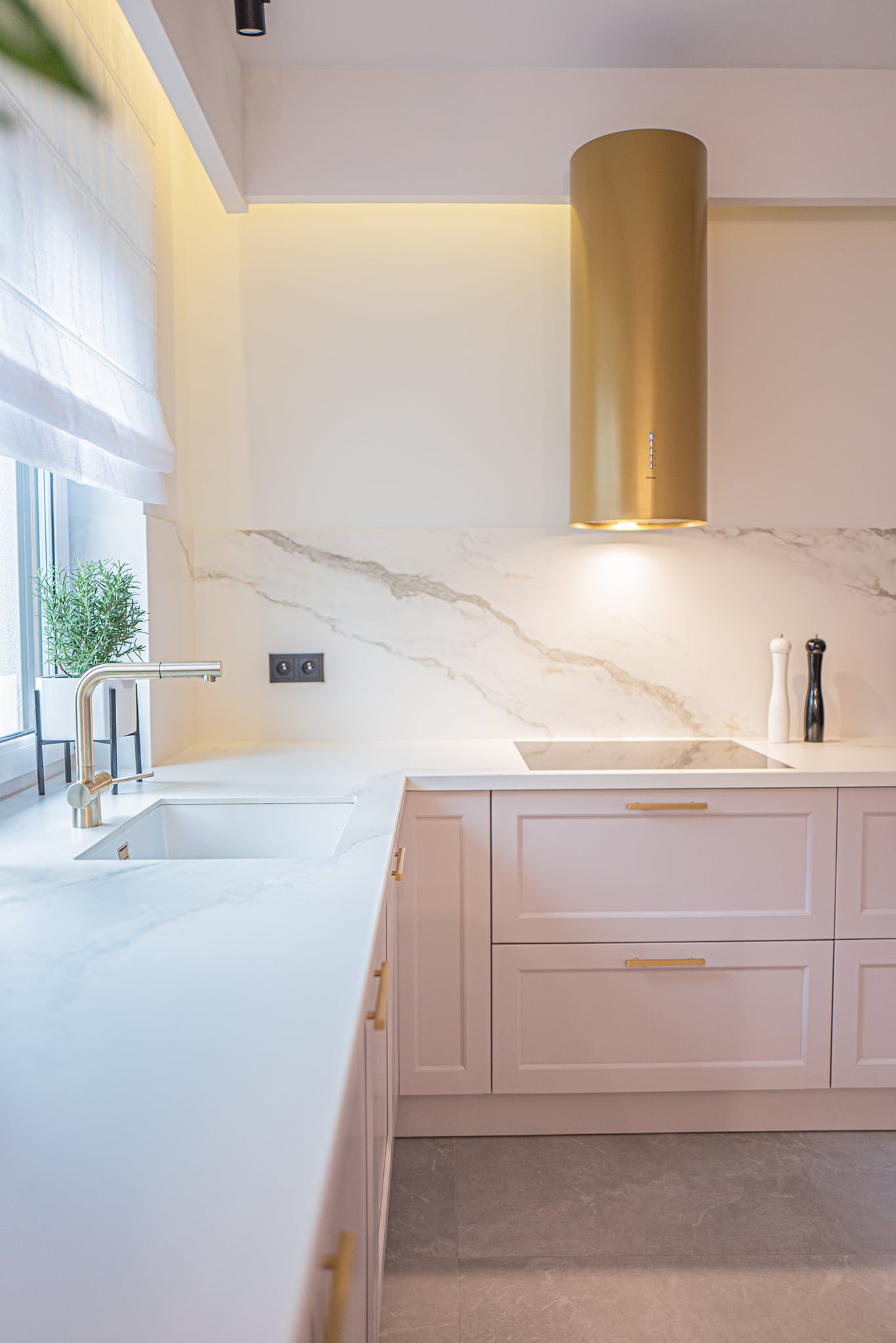 Nowoczesna kuchnia z pudrowo-różowymi szafkami, białym blatem imitującym marmur i złotym okapem cylindrycznym, uzupełniona o minimalistyczną armaturę i zioła w doniczce przy oknie.