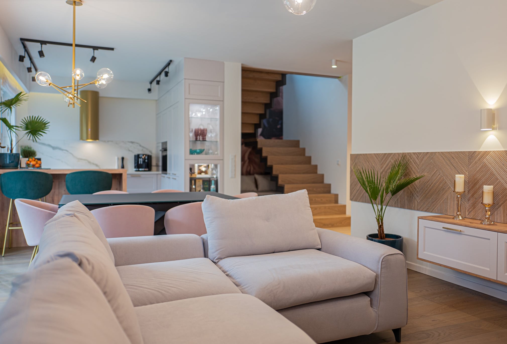 Jasnoszary narożnik z poduszkami w salonie z drewnianymi schodami w tle, widok na kuchnię z barkiem i krzesłami w odcieniach zieleni i różu, dekoracyjna ściana z drewnianą mozaiką i rośliną...