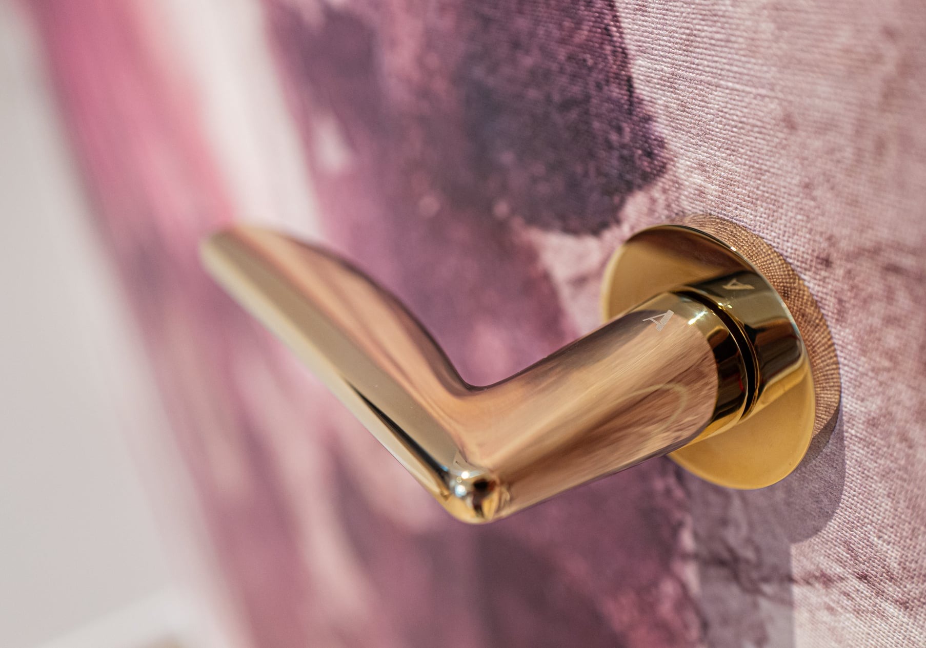 Złota klamka do drzwi z delikatnym grawerem litery 'A', zamontowana na ścianie z wzorzystą tapetą w odcieniach różu i fioletu.