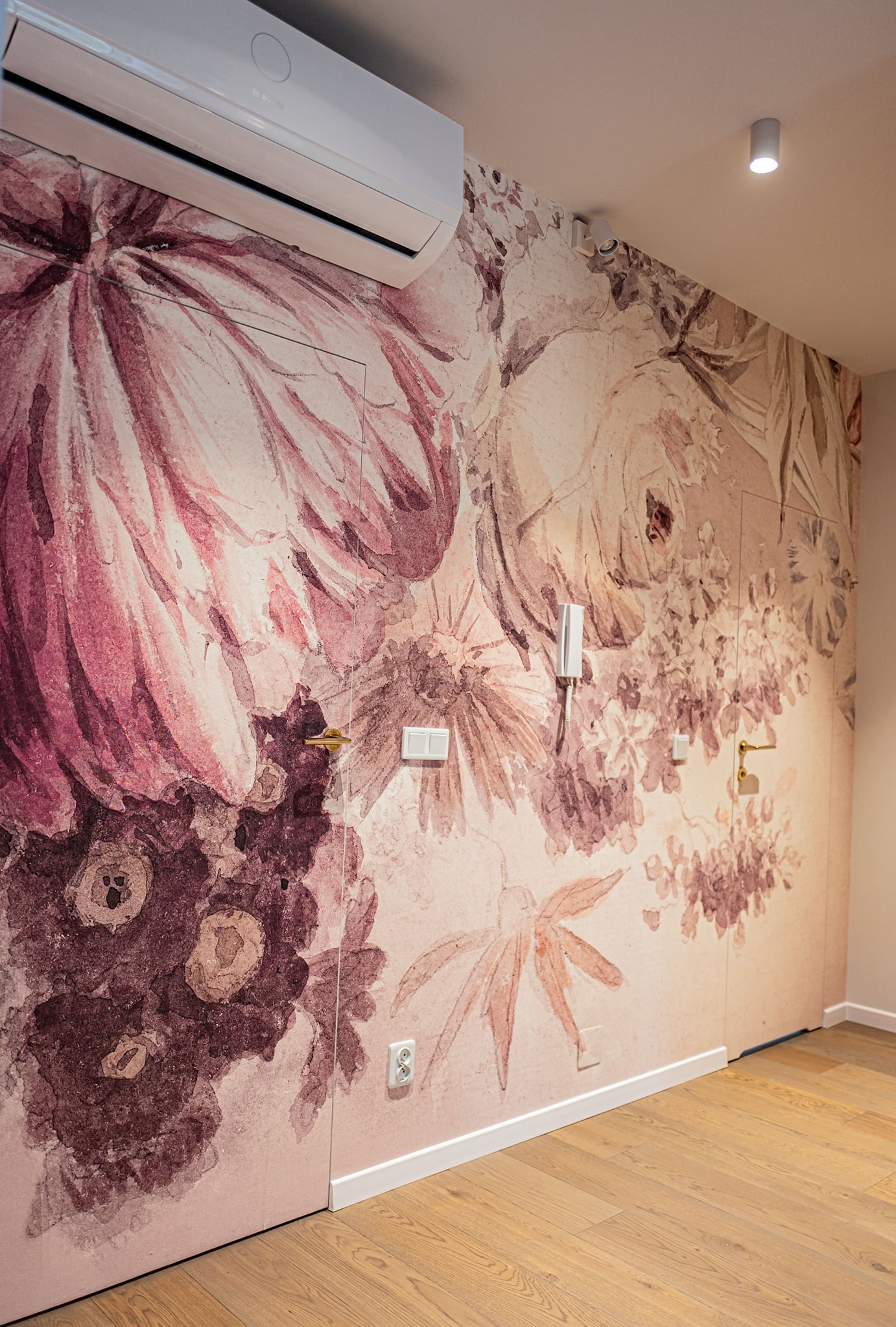 Ściana z fototapetą w kwiaty w odcieniach różu i brązu, z wkomponowanymi drzwiami i osprzętem elektrycznym, klimatyzator na górze, drewniana podłoga.