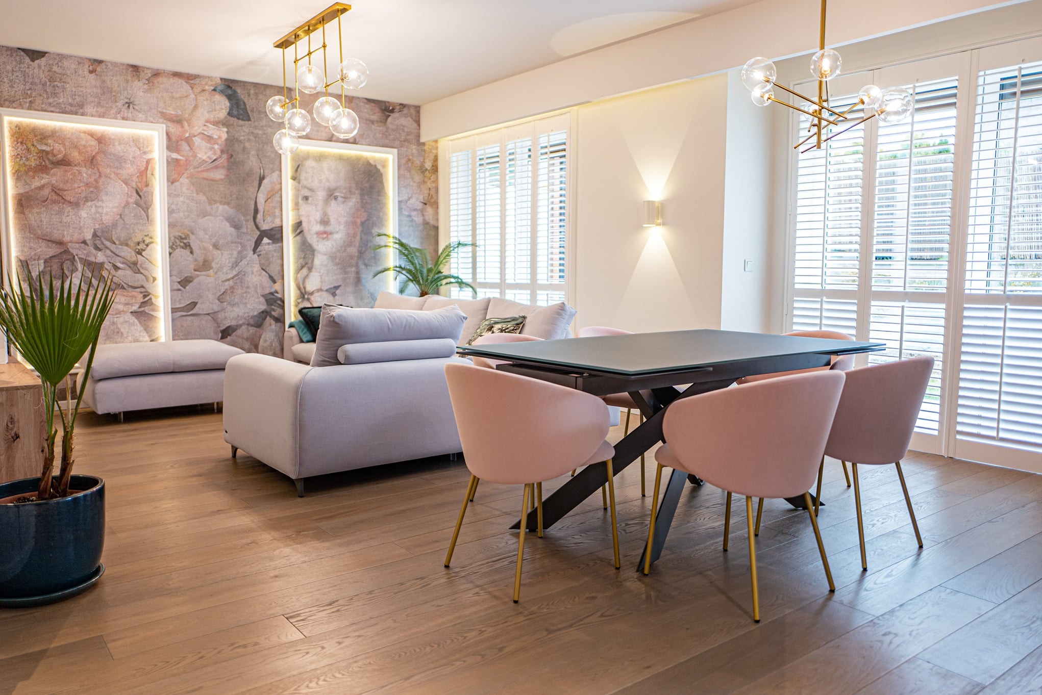 Jasne wnętrze salonu z drewnianą podłogą, szarą sofą i stołem z różowymi krzesłami na złotych nogach; na ścianie tapeta w kwiaty z portretem kobiety w ramie z podświetleniem LED.