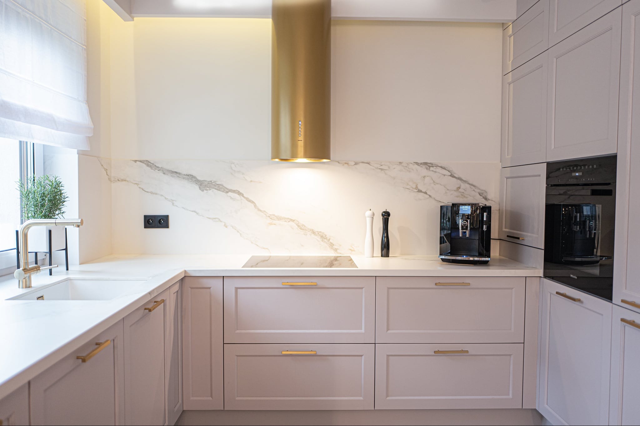 Jasna kuchnia z marmurowym backsplashiem, złotym okapem i armaturą. Na blacie ekspres do kawy i przyprawniki.