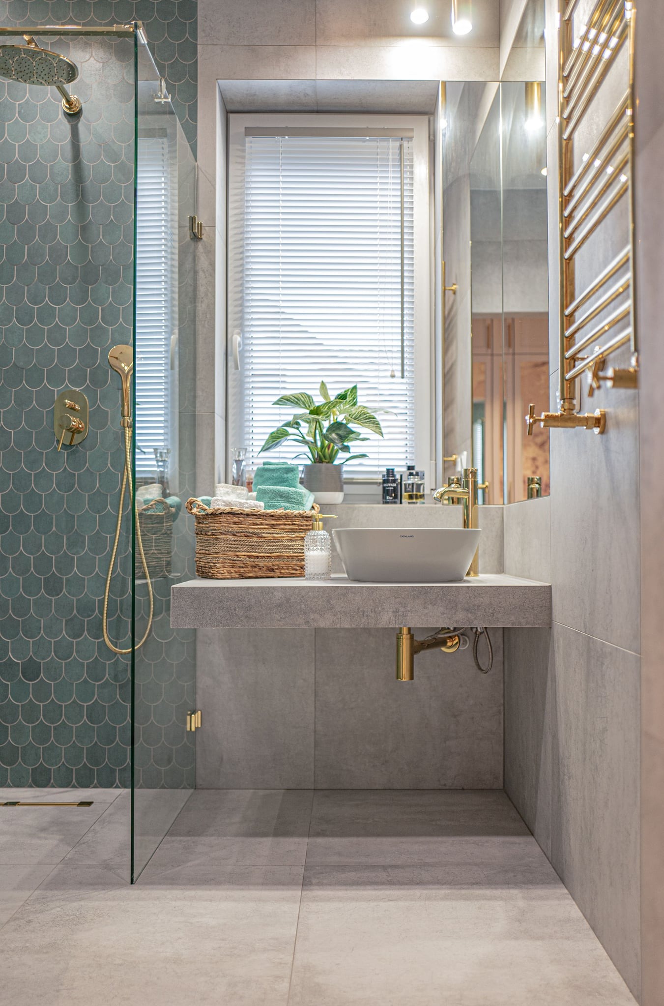 Nowoczesna łazienka z kabiną prysznicową wykończoną turkusową mozaiką w kształcie rybiej łuski, umywalką nablatową, złotą armaturą i grzejnikiem drabinkowym, uzupełniona o wiklinowy koszyk...